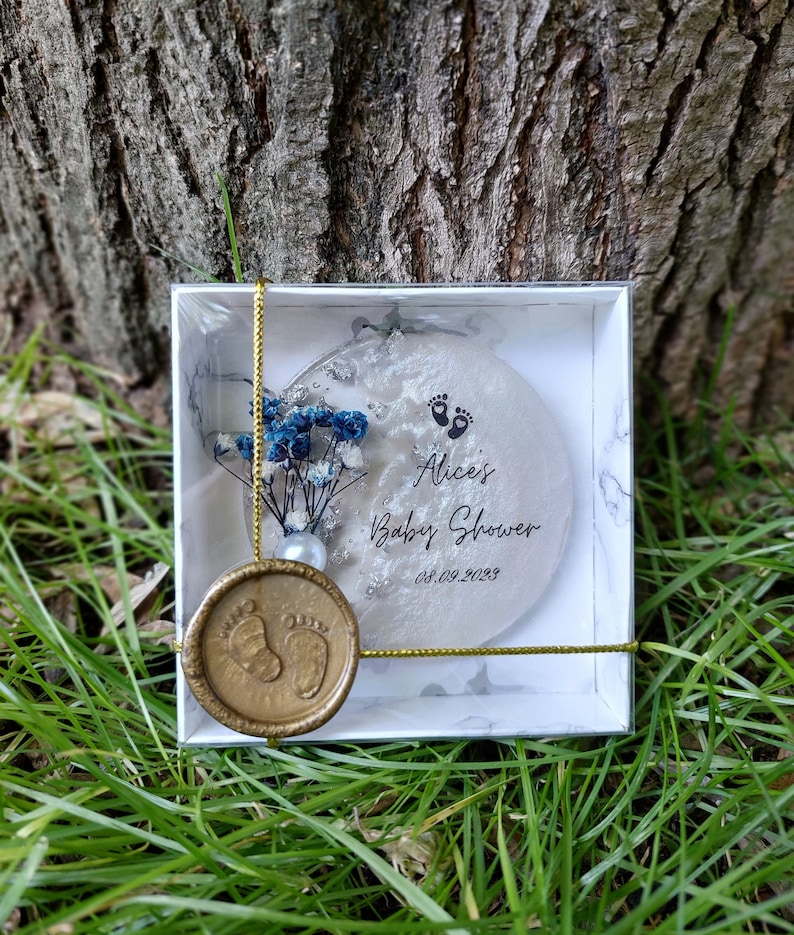 Pu&ograve; includere: Un ricordo per baby shower in una scatola bianca con un design marmorizzato. Il ricordo &egrave; un ornamento rotondo e trasparente con il testo "Alice's Baby Shower" e la data 08.09.2023. &Egrave; decorato con fiori blu e un sigillo di cera dorata.