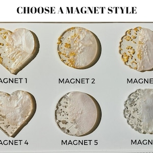 Pu&ograve; includere: Una selezione di sei calamite decorative di varie forme, tra cui cuori e cerchi. Ogni calamita presenta una base bianca perlescente con accenti di foglie d'oro o d'argento. Il testo "CHOOSE A MAGNET STYLE" &egrave; in alto.