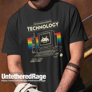 May include: A black t-shirt featuring a graphic print with the text THE MILLENNIAL'S GUIDE TO TECHNOLOGY. The design includes a vintage computer, a brick-style mobile phone, two floppy disks, a mouse, and a rainbow stripe background.