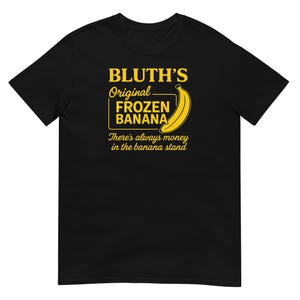 Puede incluir: Camiseta negra con texto amarillo y un gráfico de plátano. El texto dice "BLUTH'S Original FROZEN BANANA" con una ilustración de plátano. Debajo del texto está la frase "There's always money in the banana stand."