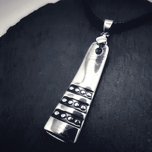 Könnte beinhalten: Eine silberne Halskette mit einem geometrischen Anhänger, der mit Reihen kleiner Kreise verziert ist. Der Anhänger ist an einem schwarzen Lederband befestigt.