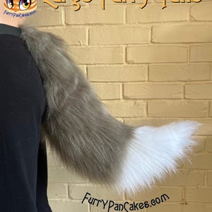 Puede incluir: Una gran cola peluda en tonos grises y blancos, de aproximadamente 84 cm de largo. La cola está sujeta a una prenda negra. El texto "Large Furry Tails" y "FurryPanCakes.com" son visibles. La imagen también indica "11 colores - Aproximadamente 33" - Cinturones incluidos."