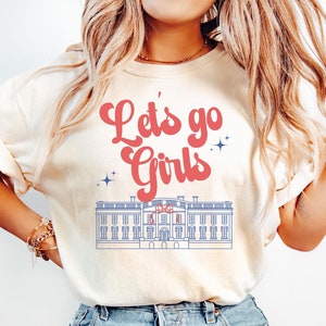 May include: A cream colored t-shirt with the text "Let's go Girls" in red and blue lettering. The shirt also features a blue and white illustration of a building.