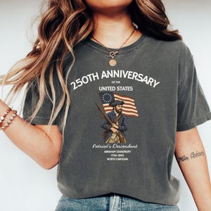 May include: Dark gray t-shirt with a graphic commemorating the 250th anniversary of the United States. The design includes an illustration of a patriot holding a rifle and the American flag, with text that reads "Patriot's Descendant".