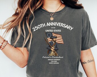 May include: Dark gray t-shirt with a graphic commemorating the 250th anniversary of the United States. The design includes an illustration of a patriot holding a rifle and the American flag, with text that reads "Patriot's Descendant".
