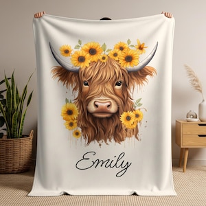 May include: A cream-colored blanket featuring a Highland cow illustration adorned with a sunflower crown. The cow has long, brown fur and large horns. The name "Emily" is written in cursive script below the cow.