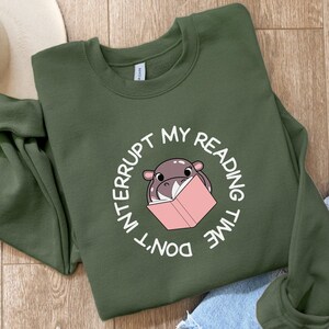 Peut inclure: Un sweat-shirt vert avec un graphique circulaire blanc représentant un hippopotame de dessin animé lisant un livre rose. Le texte autour de l'hippopotame dit "Ne pas interrompre mon temps de lecture."