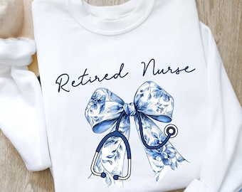 May include: White sweatshirt with the text "Retired Nurse" in blue script. A blue and white floral bow with a stethoscope is centered on the shirt.