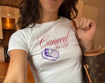 May include: White t-shirt with a red graphic that says "Canned for Life" and a blue illustration of a can of sardines. The person wearing the shirt is holding a brown leather handbag.