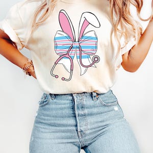 May include: A white t-shirt with a pink and blue striped bow with bunny ears and a stethoscope. The bow is centered on the shirt.