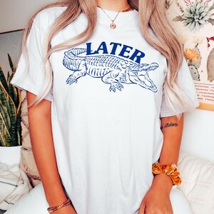 May include: White t-shirt with a blue alligator graphic and the word "LATER" printed above it.