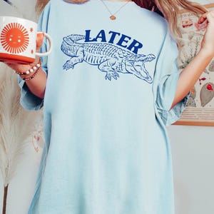May include: A light blue oversized t-shirt with a graphic of an alligator and the word "LATER" printed in blue.