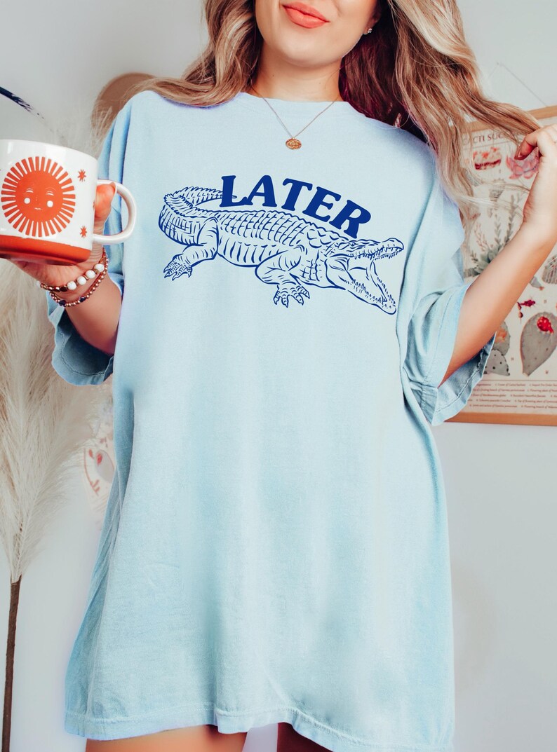 May include: A light blue oversized t-shirt with a graphic of an alligator and the word "LATER" printed in blue.