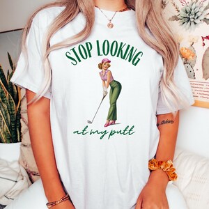 Op de afbeelding: Wit T-shirt met groene tekst die "Stop Looking at my Putt" zegt. Een cartoonafbeelding van een vrouw in een roze hoed en een groene rok die een golfclub vasthoudt, bevindt zich in het midden van het shirt.