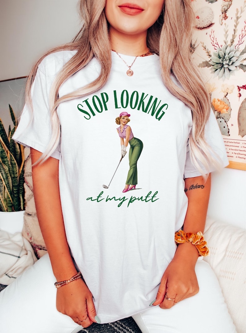 Op de afbeelding: Wit T-shirt met groene tekst die "Stop Looking at my Putt" zegt. Een cartoonafbeelding van een vrouw in een roze hoed en een groene rok die een golfclub vasthoudt, bevindt zich in het midden van het shirt.