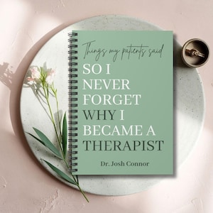 May include: A sage green spiral-bound notebook with black wire binding. The cover features the text: Things my patients said SO I NEVER FORGET WHY I BECAME A THERAPIST Dr. Josh Connor. The notebook is displayed with a small flower and a ring.