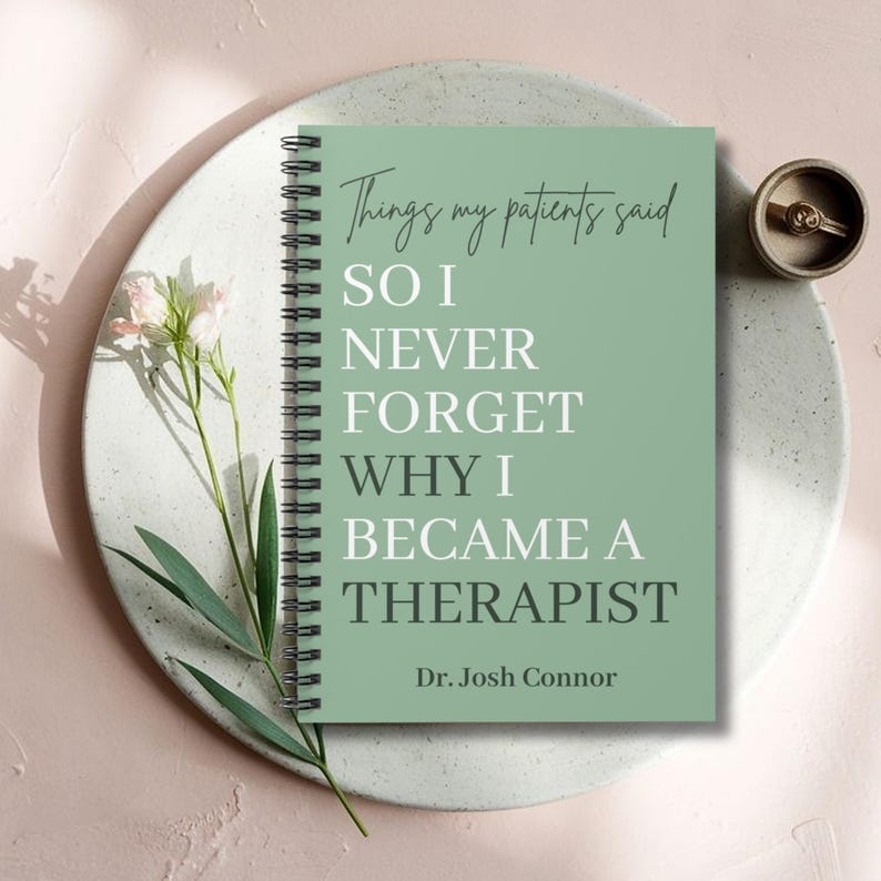 May include: A sage green spiral-bound notebook with black wire binding. The cover features the text: Things my patients said SO I NEVER FORGET WHY I BECAME A THERAPIST Dr. Josh Connor. The notebook is displayed with a small flower and a ring.