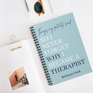May include: A spiral-bound notebook with a light blue cover featuring the text "Things my patients said SO I NEVER FORGET WHY I BECAME A THERAPIST" and "Brittany Park" in dark blue and white lettering, with a gold binder clip on the page.