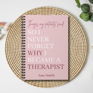 May include: A spiral-bound notebook with a dusty pink cover featuring the text "Things my patients said SO I NEVER FORGET WHY I BECAME A THERAPIST" in white and dark pink lettering, with the name "Amy Smith" printed at the bottom.