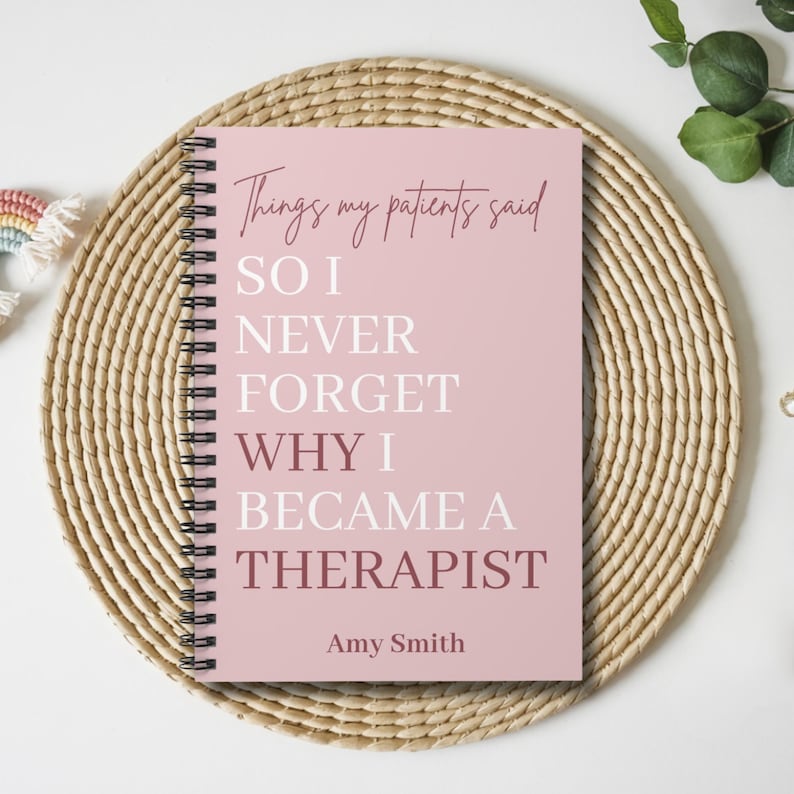 May include: A spiral-bound notebook with a dusty pink cover featuring the text "Things my patients said SO I NEVER FORGET WHY I BECAME A THERAPIST" in white and dark pink lettering, with the name "Amy Smith" printed at the bottom.