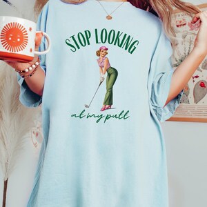 Op de afbeelding: Lichtblauw oversized T-shirt met een groene en witte afbeelding van een vrouw in een roze hoed die een golfclub vasthoudt. De tekst "STOP LOOKING at my putt" is in het groen gedrukt.