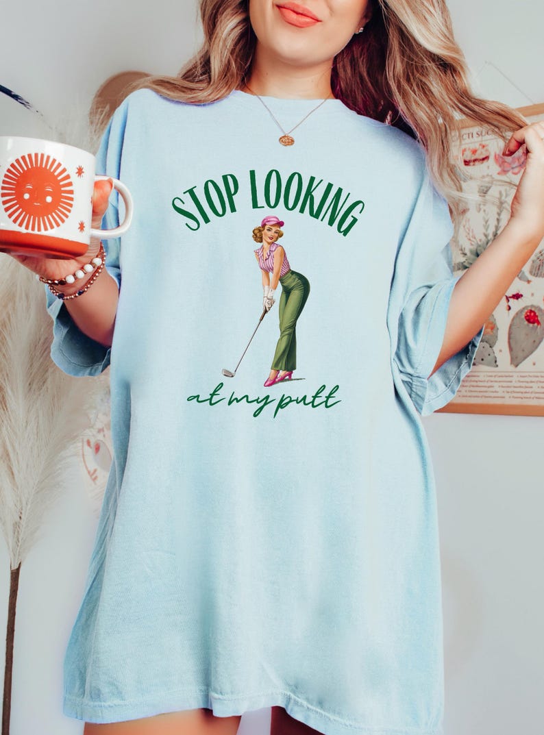 Op de afbeelding: Lichtblauw oversized T-shirt met een groene en witte afbeelding van een vrouw in een roze hoed die een golfclub vasthoudt. De tekst "STOP LOOKING at my putt" is in het groen gedrukt.