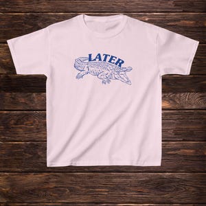 May include: A white t-shirt featuring a blue alligator graphic with the word "LATER" printed above it.