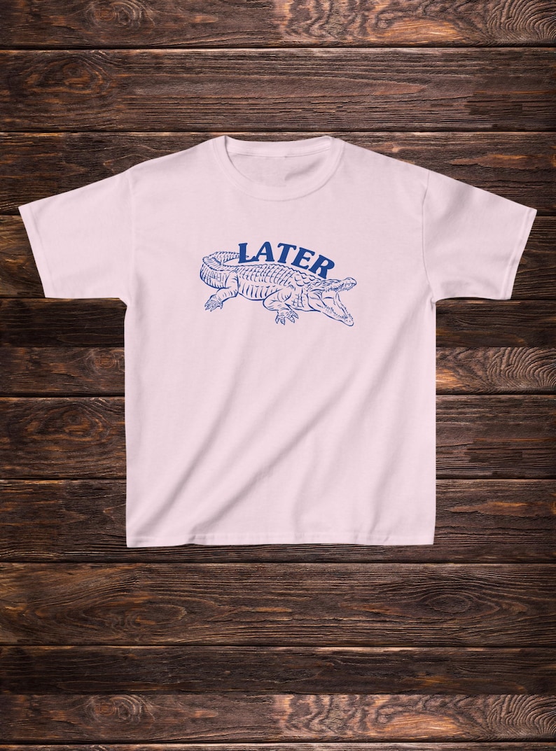 May include: A white t-shirt featuring a blue alligator graphic with the word "LATER" printed above it.