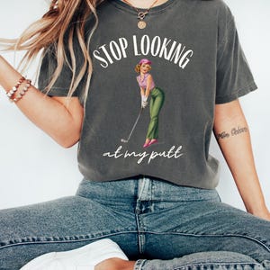 Op de afbeelding: Een grijs T-shirt met de tekst "STOP LOOKING at my putt" en een vintage illustratie van een vrouw die golf speelt.