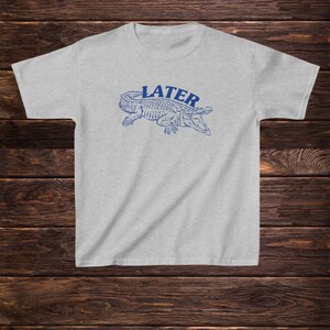 May include: A light grey t-shirt with a blue alligator graphic and the word "LATER" printed above it.