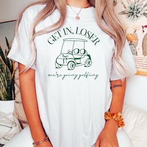 May include: White t-shirt with green text that says "Get in, Loser we're going golfing" and a green illustration of a golf cart.