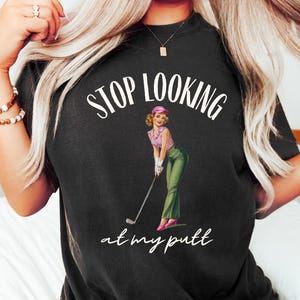 Op de afbeelding: Een zwart T-shirt met de tekst "STOP LOOKING at my putt" in wit. In het midden van het shirt staat een cartoonafbeelding van een vrouw in een roze hoed en een groene rok die een golfclub vasthoudt.