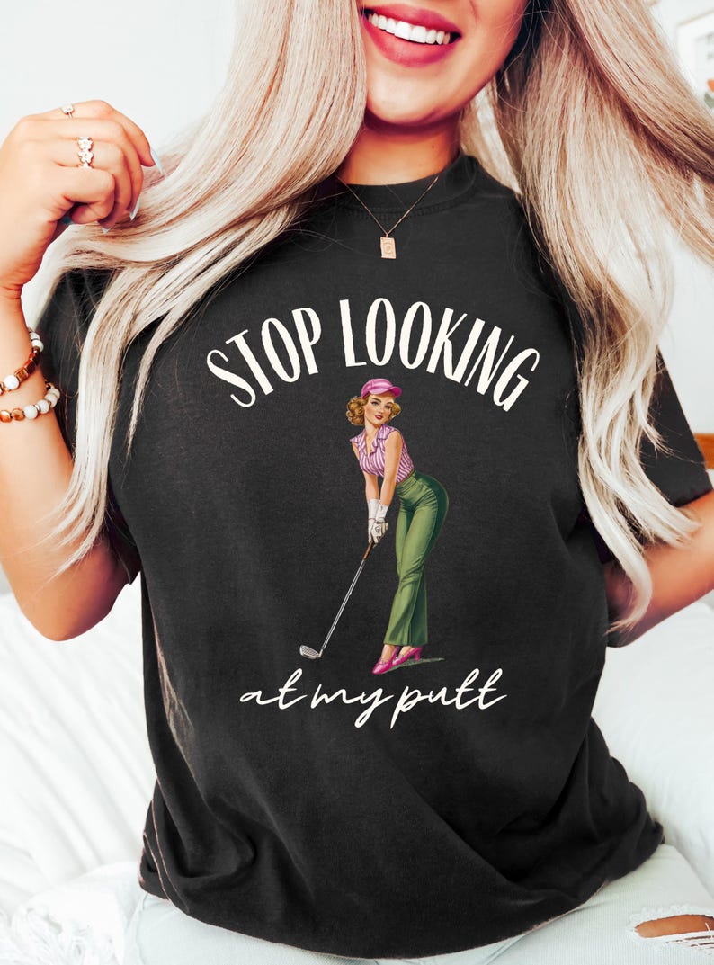 Op de afbeelding: Een zwart T-shirt met de tekst "STOP LOOKING at my putt" in wit. In het midden van het shirt staat een cartoonafbeelding van een vrouw in een roze hoed en een groene rok die een golfclub vasthoudt.