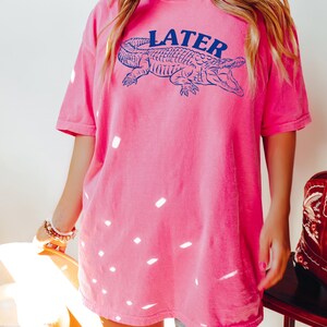 May include: A pink t-shirt with a blue alligator graphic and the word "LATER" printed on it.