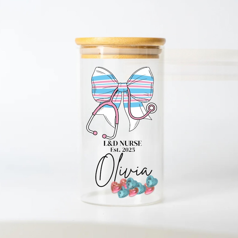 May include: A clear glass jar with a bamboo lid, decorated with a pink and blue striped bow and a pink stethoscope. The jar has the text "L&D NURSE Est. 2025 Olivia" printed on it. The jar is filled with small pink and blue heart-shaped beads.