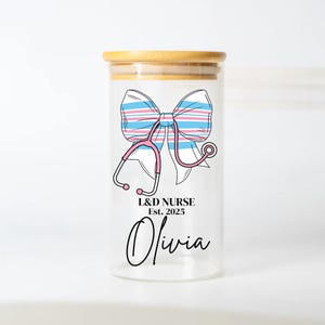 May include: A clear glass jar with a bamboo lid, featuring a pink stethoscope and a blue and pink striped bow. The jar has the text "L&D NURSE Est. 2025 Olivia" printed on it.