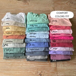 May include: A collection of 24 t-shirts in various colors, stacked in two rows. The colors are labeled with names like "white", "wory", "butter", "island reef", "chalky mint", "seafoam", "hydrangea", "chambray", "blue jean", "flo blue", "washed denim", "denim", "gray", "pepper", "peachy", "crunch berry", "blossom", "watermelon", "heliconin", "brick", "chili", "orchid", "violet", "berry", and "black". The text "COMFORT COLORS 1717" is displayed at the top of the image.