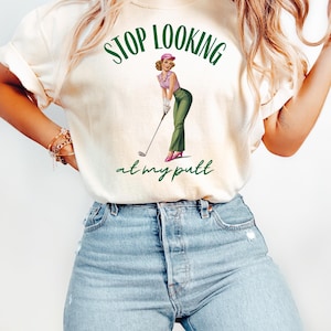 Op de afbeelding: Een wit T-shirt met de tekst "STOP LOOKING at my putt" in groene letters. Boven de tekst staat een cartoonafbeelding van een vrouw in een roze hoed en een groene broek die een golfclub vasthoudt.