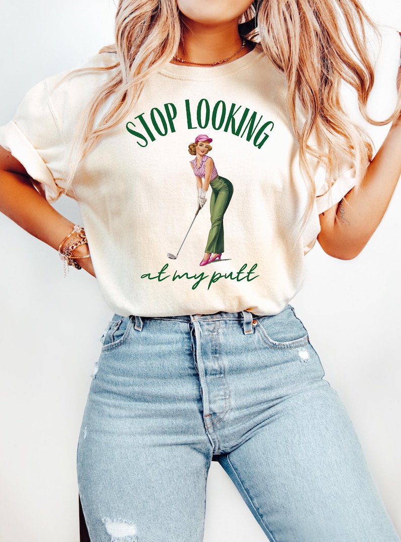Op de afbeelding: Een wit T-shirt met de tekst "STOP LOOKING at my putt" in groene letters. Boven de tekst staat een cartoonafbeelding van een vrouw in een roze hoed en een groene broek die een golfclub vasthoudt.