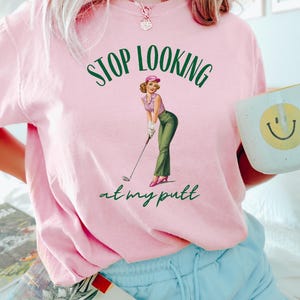 Op de afbeelding: Roze T-shirt met een groene afbeelding van een vrouw die golf speelt. De afbeelding zegt "STOP LOOKING at my putt".