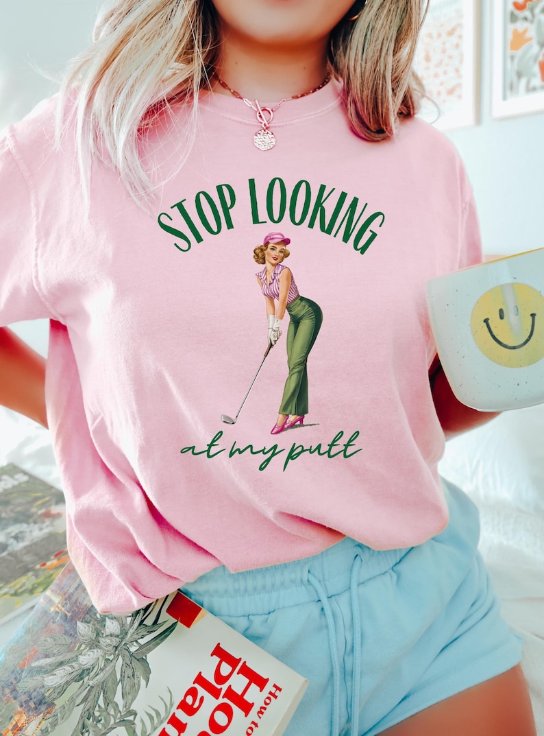 Op de afbeelding: Roze T-shirt met een groene afbeelding van een vrouw die golf speelt. De afbeelding zegt "STOP LOOKING at my putt".