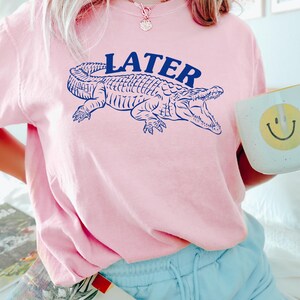 May include: A pink t-shirt with a blue alligator graphic and the word "LATER" printed on the front. The shirt is being worn by a person.