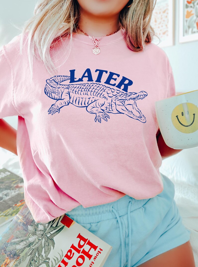 May include: A pink t-shirt with a blue alligator graphic and the word "LATER" printed on the front. The shirt is being worn by a person.