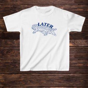 May include: White t-shirt featuring a blue alligator graphic with the word "LATER" above it.