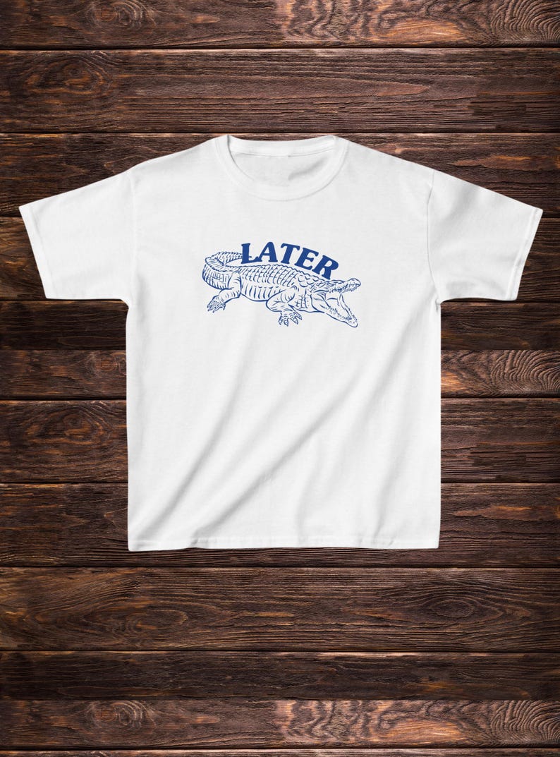 May include: White t-shirt featuring a blue alligator graphic with the word "LATER" above it.