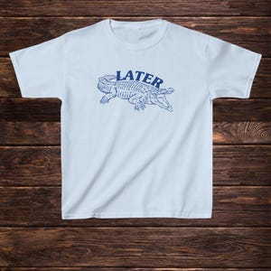 May include: Light blue t-shirt featuring a blue alligator graphic and the word "LATER" printed on the front.