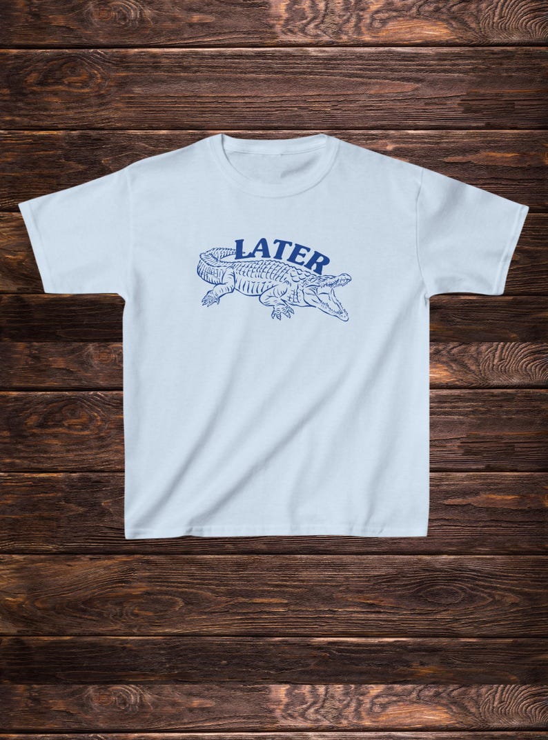 May include: Light blue t-shirt featuring a blue alligator graphic and the word "LATER" printed on the front.
