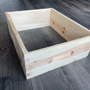 May include: A wooden beehive box with a natural finish. The box has a rectangular shape and is made of light-colored wood. The box has a small opening on one side.