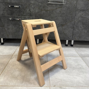 Puede incluir: Un taburete de madera con dos peldaños. El taburete está hecho de madera clara y tiene un diseño sencillo.