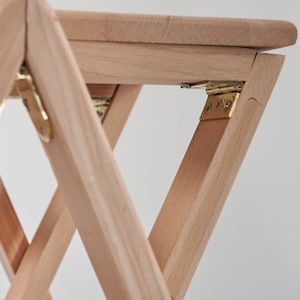 May include: A wooden folding stool with brass hinges. The stool is made of light wood and has a simple design.